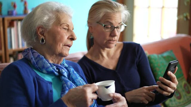 Elderly senior woman and mature daughter drink tea looking at smartphone in living room.