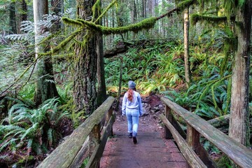 A young woman walking along a trail in the beautiful evergreen forests outside Vancouver, British Columbia, Canada.  She is walking along a trailwith large trees, moss, ferns -- very beautiful