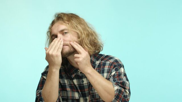 Mirror reflection of handsome blond man with beard looking at his face, pop pimples and zits, squeezing acne, standing over blue background