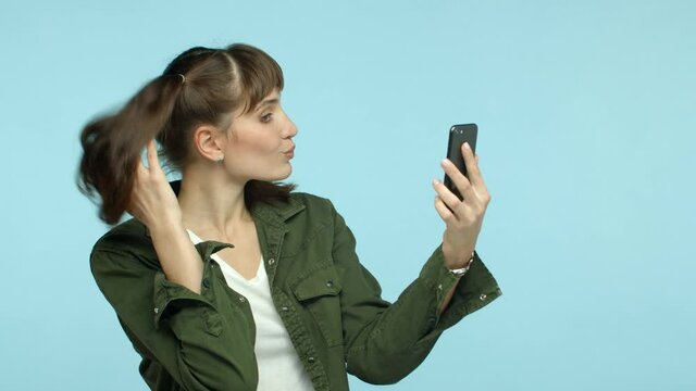 Slow motion of attractive sassy woman with double ponytail hairstyle, taking selfies with photo filters, posing and making cheeky face, looking at smartphone camera, turquoise background