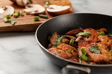 Side view selective focus image of sauteed roasted chicken breasts in a non stick frying pan on white marble seasoned with parsley, spring onion and mushroom as well as herbs, tomato sauce and spices.