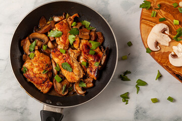Flat lay image of a non stick frying pan with a chicken breast sautee inside. Meat is seasoned with herbs, tomato, spices, parsley, spring onions and mushrooms. There is a chopping board