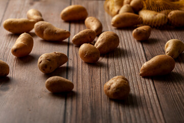Shallow depth of field dark image of small irregular shaped ratte potatoes scattered on the surface of a wooden table from a tipped over mesh sack. An artistic versatile low light still life photo.