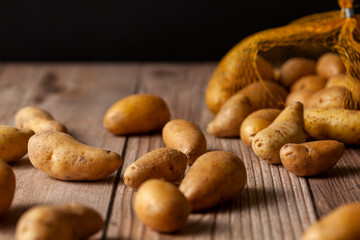 Shallow depth of field dark image of small irregular shaped ratte potatoes scattered on the surface of a wooden table from a tipped over mesh sack. An artistic versatile low light still life photo.