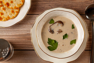 Flat lay image of Creamy mushroom soup decorated with parsley leaves in an artisan porcelain cup on a flat plate on a wooden table served with crackers,  spoon and a salt shaker.
