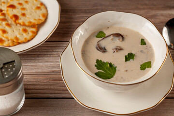 Side view of Creamy mushroom soup decorated with parsley leaves is in an artisan porcelain cup on a flat plate on a wooden table served with crackers,  spoon and a salt shaker. An upscale vintage
