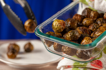 Close up image of a thick glass bowl of cooked and seasoned Brussel sprouts being held with kitchen cloth and served into porcelain plates using a tong. Healthy, simple, vegan, homemade eating concept