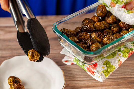 Close Up Isolated Image Of A Thick Glass Bowl Of Cooked And Seasoned Brussel Sprouts Being Held With Kitchen Cloth And Served Into Porcelain Plate On Wooden Table Using A Tong. Vegan Homemade Eating.