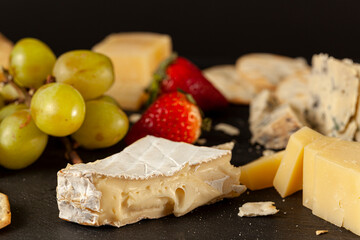 A selection of cave aged artisan French, Italian and Swiss cheese assortment on black cheese board served with crackers and fruits (grapes and strawberries) . A dark background close up image.