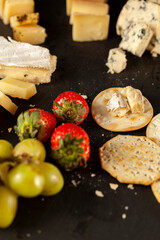 A vertical image of a cheese platter containing a selection of aged artisan French, Italian and Swiss cheese assortment on black cheese board served with crackers, strawberries and grapes.