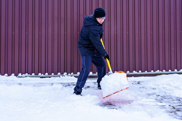 An elderly European, 61, shovels snow from the gate with an orange shovel. Healthy lifestyle...