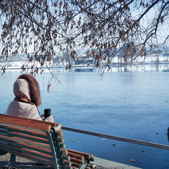 A girl in a winter coat was sitting on a bench near an artificial lake, looking at the frozen lake and drinking hot tea from a thermos.