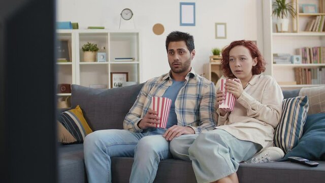 Medium long of young Mixed-Race man sitting on couch together with ginger-haired Caucasian girlfriend, watching movie, getting scared, scattering popcorn