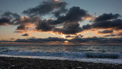 Obraz premium Sunset. The sun's rays make their way through the dark blue clouds, illuminating them with orange. Sea waves roll with foam and spray onto the pebble beach. Black Sea.