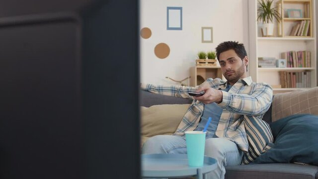 Tracking right shot of Mixed-Race guy sitting on couch at home, switching channels, ginger-haired girlfriend bringing bowl of popcorn, hugging boyfriend from back