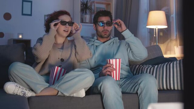 Steadicam of Mixed-Race young man sitting next to red-haired Caucasian woman in lotus pose on couch in apartment, holding popcorn boxes, impressed by looking through 3D glasses on TV