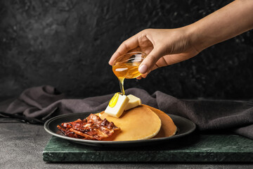 Woman pouring honey onto tasty pancakes with fried bacon and butter on table