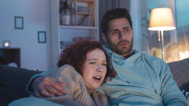 Tracking right of dark-haired young man in blue hoodie sitting on couch at home, Caucasian girlfriend lying in his arms, couple watching horrible movie scene on invisible TV