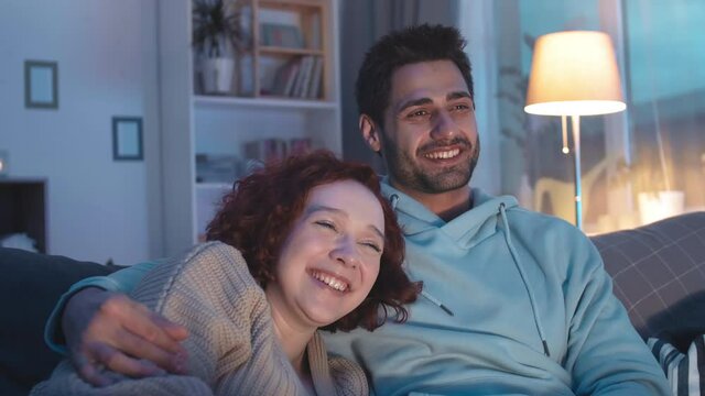 Close-up of multiethnic romantic partners snuggling on sofa in apartment at night, watching invisible TV, laughing, Mixed-Race young man hugging Caucasian girlfriend