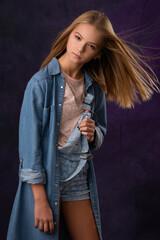 beautiful girl blonde teenager in denim overalls and denim unbuttoned raincoat posing on a dark background