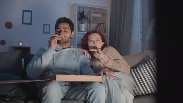 Medium long of young Mixed-Race guy and red-haired Caucasian girl sitting in apartment on couch, cringing, eating pizza from box, watching horror movie on invisible TV, then laughing