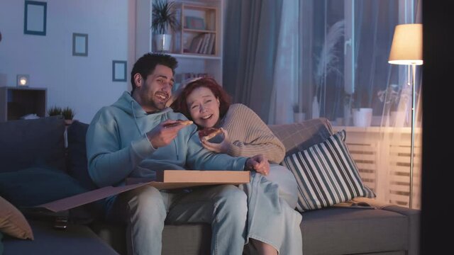 Medium long steadicam of young Mixed-Race man eating pizza from box, his Caucasian girlfriend sitting near on couch in living room. Couple cringing, talking and laughing at invisible movie scene