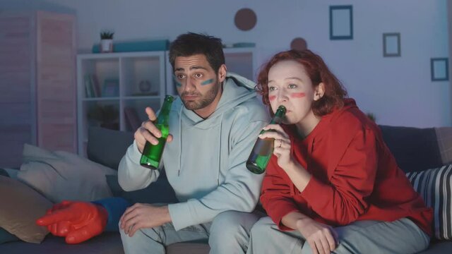 Tracking right medium long of diverse sport fans with football stripes painted on faces sitting on couch at night, watching game on TV, drinking bottled beer, talking. Woman in red hoodie, man in blue