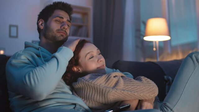 Steadicam shot of young romantic couple watching TV at night at home on couch. Loving Mixed-Race man hugging, kissing on head, running hand through hair of happy Caucasian girlfriend