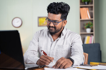 Fototapeta na wymiar Young man writing down notes by looking laptop - concept of employee or student online training at home.