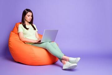 Photo portrait full body view of concentrated woman sitting in bean bag with laptop isolated on vivid violet colored background