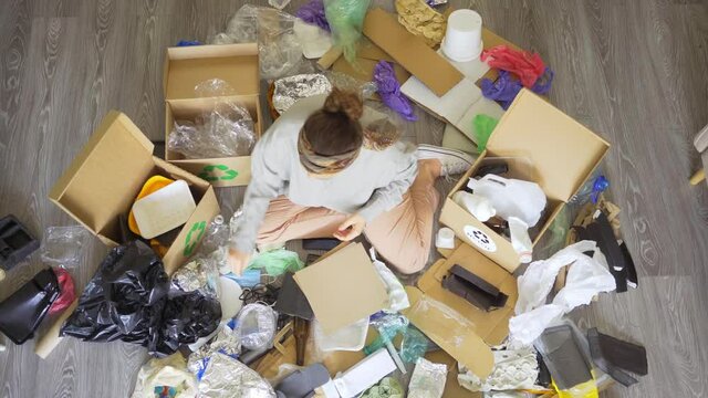 Above view faceless woman sorting different kinds of waste while sitting on floor among pile of garbage and cardboard boxes in light living room, footage with circle moving 