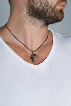Cropped shot of bearded man wearing black rubber necklace cord with silver pendant in the form of wolf head. The man in white t-shirt is posing on the gray background. 