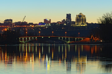 Naklejka premium Scenic night cityscape of Kyiv?s hills with modern skyscrapers. Citylights reflected in tranquil water of Dnipro River. Bridge and bare trees in the Hydropark