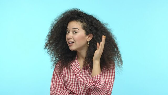 Slow motion of sassy girl with long curly hair cant hear you, looking confused with hand near ear, dont understand what saying, standing over blue background