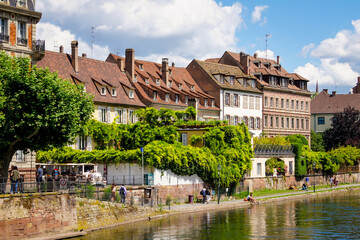 Beautiful view of a riverfront with houses and scenic gardens full of fresh green wisteria. Peope are sitting at the riverfront and enjoy a warm summer day.