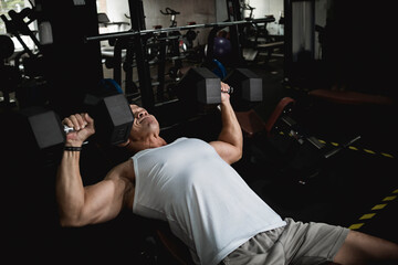 A fit asian guy in a white tank top does some incline dumbbell presses. Working out at the gym,...