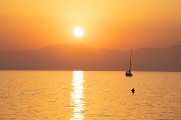 Aurora con sole nascente sulla montagna , riflesso sul mare e barca a vela . Tonalit&agrave; generale colore molto calda