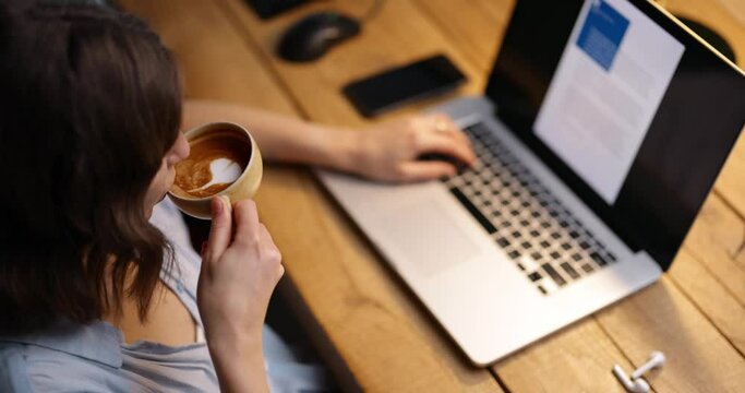 Female working on laptop and drinking coffee with a heart shape. High angle, close-up tracking shot.
