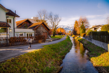 Happy girl in Alps mountains in Liechtenstein. Medieval Red House, calm narrow mountain river and jogging track, residential buildings, blue sky and snow-capped mountains. Liechtenstein, Vaduz