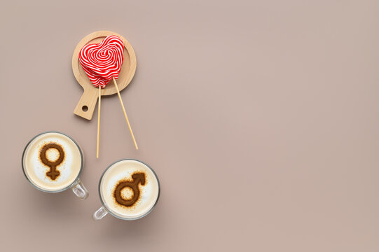 Two cups of coffee with symbols of mars and venus on whipped milk foam and couple of lollipops in heart shape on beige tabletop. Concept romantic date on Valentine's day. Flat lay