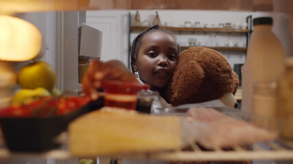 Cute african kid holding teddy bear taking yogurt from fridge