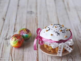 Easter bun decorated with cream with pastry sprinkles and a ribbon bow, with colored eggs on a light wooden background. Pastry treat.