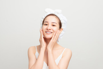 Young girl with fresh and clean skin, wearing white bath towel on her head, touching her face, looking with cute smile against blank wall