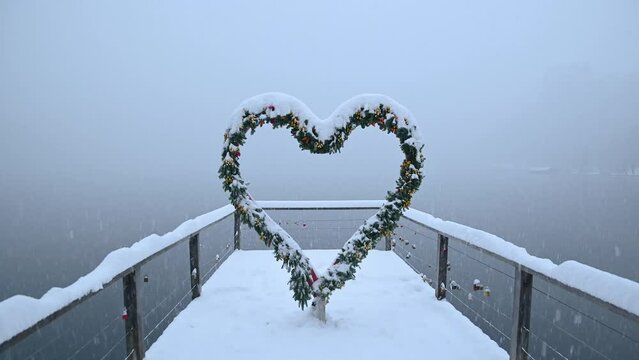 Snowstorm in winter season. Lake Bled with heart shaped stand on wooden pier covered with snow. Christmas decoration. Famous picture spot for lovers. Static shot, real time, wide angle