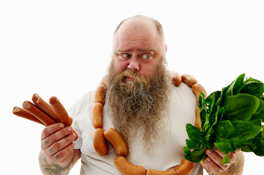 An Overweigh Man Choosing What Is Better For Health :Sausages Or Greens. Bearded Obese Man With A Bruch Of Sausages Around The Neck.