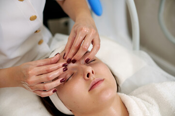 Hands of beautician making facial lifting massage for pretty young woman in SPA salon