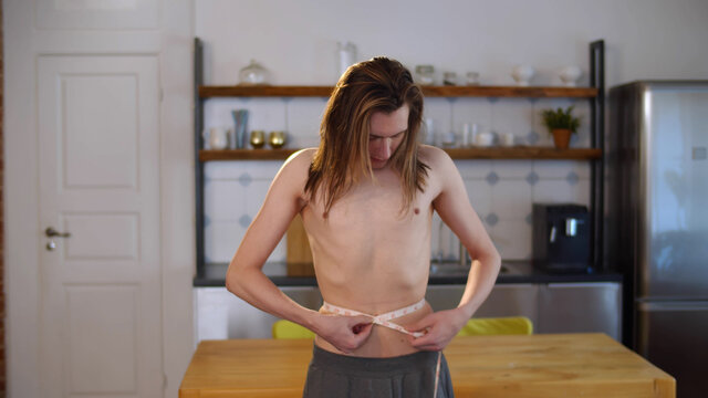 Young thin shirtless man measuring waist with tape at home