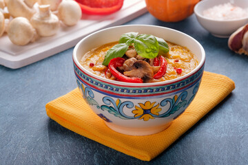 A bowl of pumpkin cream soup with basil, roasted mushrooms and red bell pepper.