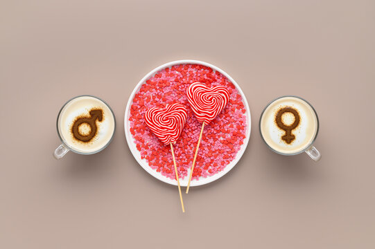 Two cups of coffee with symbols of male and female on whipped milk foam and lollipops in heart shape on white plate. Beige background. Concept romantic date on Valentine's day. Flat lay