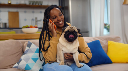 Portrait of beautiful young afro woman stroking dog and speaking on mobile phone at home